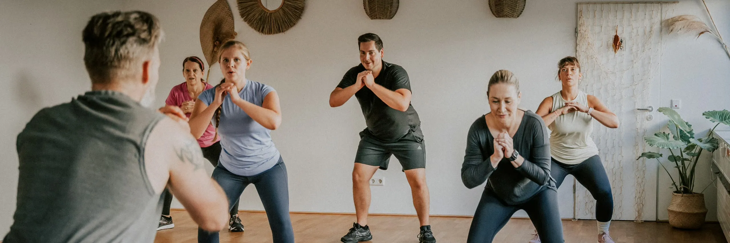 Eine Kursgruppe hält gemeinsam die Kniebeuge, der Trainer macht sie vor