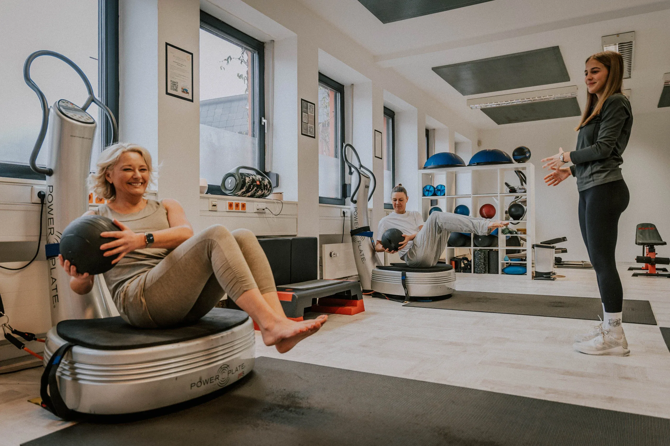 Zwei Frauen trainieren lachend mit Medizinbällen auf Vibrationsplatten, eine Trainerin leitet an