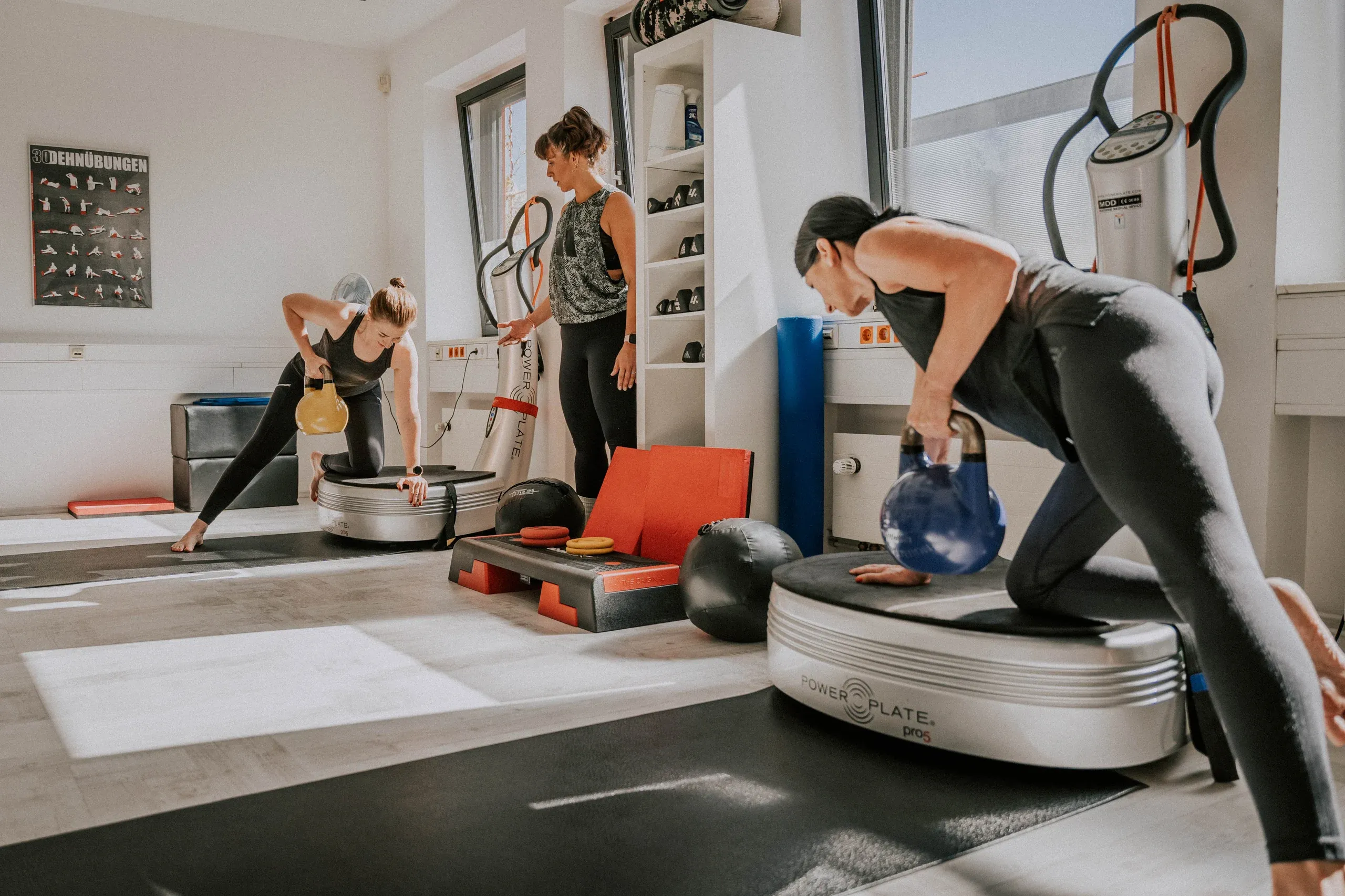 Zwei Frauen stützen sich mit Kettlebells auf ihren Vibrationsplatten ab, eine Trainerin steht daneben