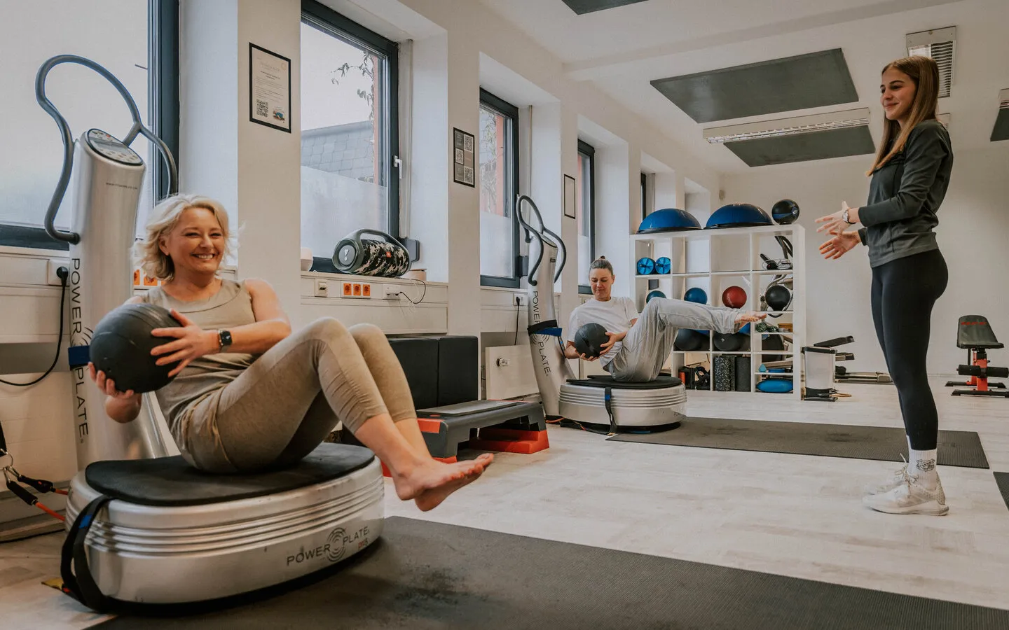 Zwei Frauen trainieren mit Medizinbällen auf Vibrationsplatten, eine Trainerin steht daneben und leitet an